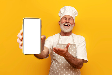 cheerful old grandfather chef in apron and hat shows blank smartphone screen on yellow isolated...