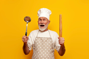 shocked old grandfather chef in apron and hat is surprised and holds dishes on yellow isolated background