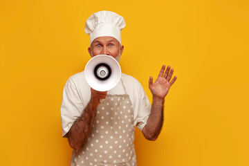 old grandfather chef in apron and hat announces information into megaphone on yellow isolated background