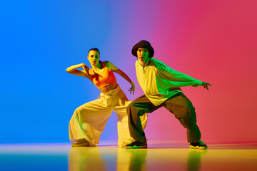 Artistic, talented, expressive young woman and man, dancers performing hip hop against blue red background in neon light. Concept of hobby, action, street style, contemporary dance, youth, fashion