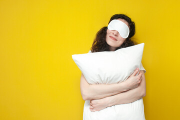 woman in sleep mask hugs white soft pillow on yellow background and sleeps, the concept of healthy...