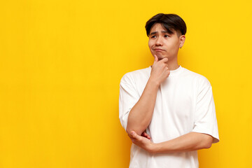 pensive young asian guy in white t-shirt thinks and plans on yellow isolated background