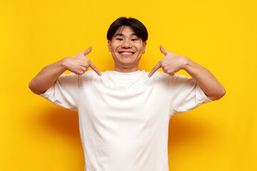 proud young asian guy in white t-shirt smiling and pointing to himself over yellow isolated...