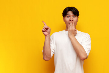 shocked young asian guy in a white t-shirt points with his hand to the side on a yellow isolated...