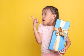 Excited Asian little girl holding a gift box and pointing aside to the empty space