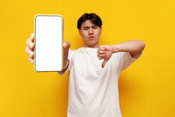 young guy asian student shows blank smartphone screen and dislike on yellow isolated background