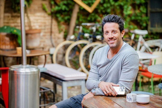 Portrait of a handsome young man sitting at bar table on terrace, looking at camera with a broadly smile - brewery pub terrace