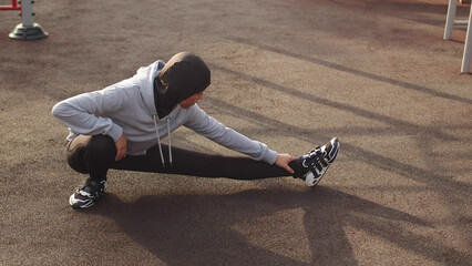 Young Asian Muslim Sportswoman In Hijab Alternately Stretching Her Legs Outdoor