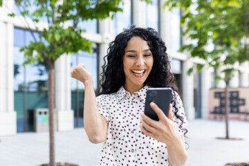 Young joyful woman winner received online notification on phone, Hispanic woman with curly hair celebrating success and triumph walking in city near office building outside - obrazy, fototapety, plakaty