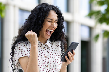 Young joyful woman winner received online notification on phone, Hispanic woman with curly hair celebrating success and triumph walking in city near office building outside