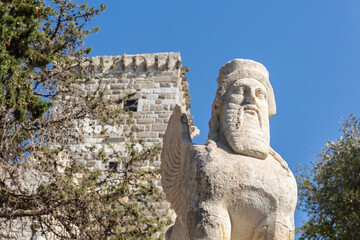Fototapeta na wymiar Statue of the ancient winged sphinx with the head of a bearded man and the medieval tower and blue sky at background. Bodrum (Mugla), Turkey (Turkiye)
