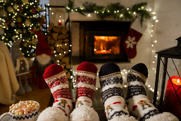 Couple enjoying by warm fireplace and warming up their feet in woolen socks with Christmas ornaments and Christmas tree and decorations background. Cozy Winter, Christmas and Family.