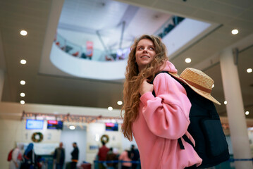 Joyous woman with backpack and hat anticipates holiday journey inside busy airport adorned for...