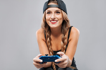 Cheerful young woman in hat and pigtail hairstyle playing console with joystick on gray background...