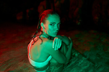 Woman immerses in lagoon, nocturnal setting with red and green illumination. Reflective mood,...