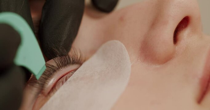 Closeup on beautician in gloves doing lash lamination procedure to woman client in beauty salon. Master with applicator instrument make eyelash care, lifting and curling treatment to young female