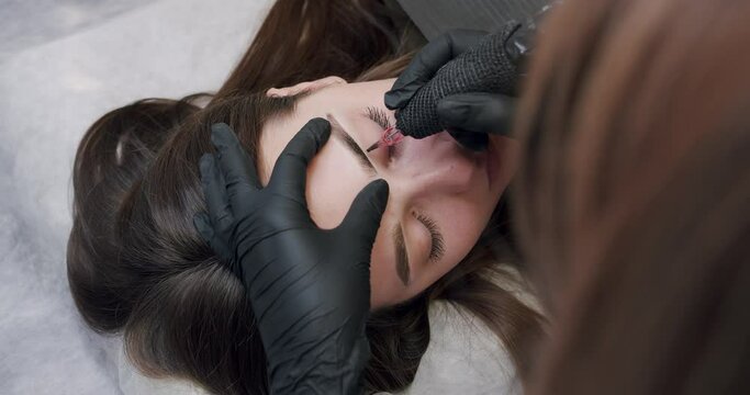 Beautician in black gloves making permanent makeup of eyebrows to young client woman in beauty salon. Cosmetologist female with professional tool doing microblading procedure, brow tattoo. 