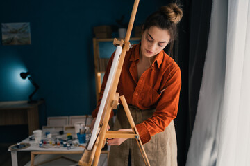 Female artist in an apron in a home studio stands near an easel and looks on it. hobby and vacation concept.