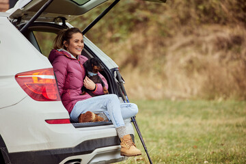 A smiling female dog owner and her dog, sitting in the car trunk, being outdoors.