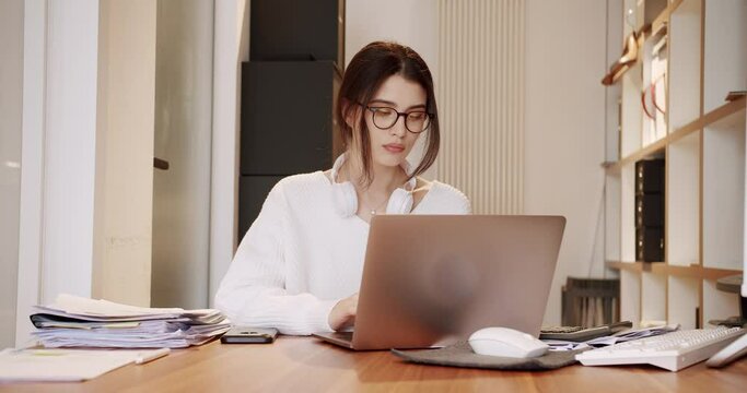 Focused pretty woman accountant in eyeglasses sitting at desk and working on laptop in office. Serious female typing on computer and enter data. Concentrated bookkeeper at comfort workplace.