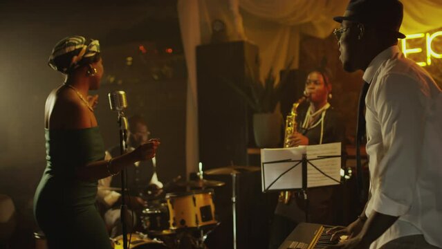 Medium shot of African American jazz band playing musical instruments and female vocalist singing while giving concert on stage in retro jazz club at night