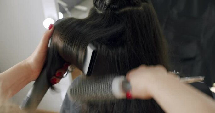 Hairdresser doing hairstyle with brush and hair dryer to woman client at beauty salon. Hairstylist drying brunette female hair and making styling using hairbrush