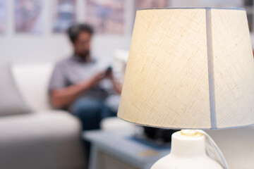 Close up of lamp spending warm light at cozy reader's corner with blurred of man sit on sofa behind.