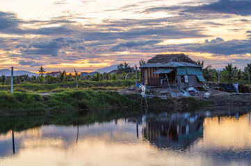Obraz premium An old shack, located by a river in central Vietnam. The sun is setting through a cloudy sky.