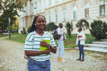 Portrait of a young woman carrying her schoolbooks outside at college. African American college girl looking at the camera with a smile. Student Standing at Entrance to University Building
