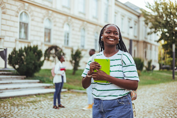 Fototapeta premium Cheerful African American Female Student With Smartphone And Workbooks Standing Outdoors, Happy Woman Walking In City After College Classes, Looking Away And Smiling, Copy Space