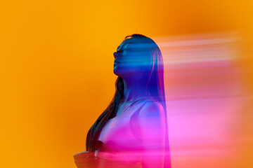 Portrait of young woman posing against orange studio background with neon colored reflection on...