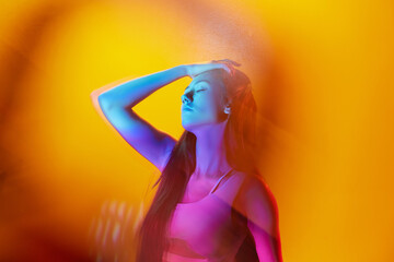 Portrait of young woman posing against orange studio background with neon colored reflection on...