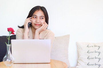 happy asian woman sitting on sofa answer call on smartphone and working with laptop wearing casual clothes, female designer working on project at home