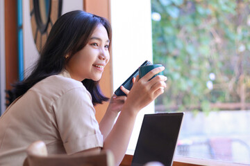 happy asian woman sit in cafe drink a cup of coffe and using smartphone with laptop on table wearing fashionable clothes, beautiful female work from coffee shop, relaxation