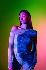 Portrait of young beautiful girl posing against gradient pink green background with neon...