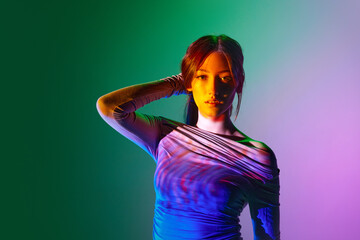 Beautiful young woman posing against gradient pink green background with neon reflection on face...