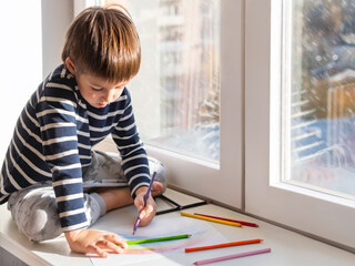 Left-handed boy sits on window sill and draws rainbow with colored pencils. Creative leisure activity for children at home.