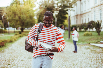 Portrait of a young African American male student smiling and carrying books and a backpack, going...