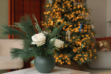 Roses, evergreen branches in vase on table, illuminated Christmas tree on background. Stylish living room decorated with Christmas ornaments and decorations. 