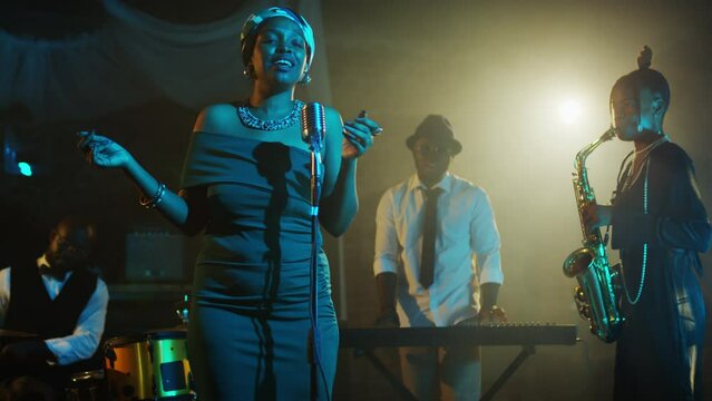 Medium shot of young African American female jazz singer performing lyric song in retro mic while giving live concert together with music band playing instruments beside
