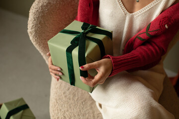 Close-up of a stylish woman holding a gift box decorated with a green ribbon at home.