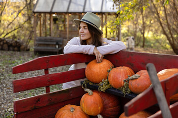 Smiling woman in a hat on a farm. Pumpkin harvest in a wooden cart.