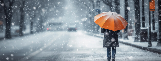 Girl walking on cold day in winter through a road with orange color umbrella and surrounded by...
