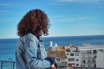 chica mira al mar que esta azul, en un mirador, por encima de unos tejados
