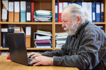 Elderly man with laptop at workplace in office, side view