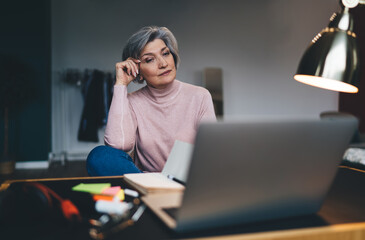 Focused mature woman working on laptop and taking notes in notebook