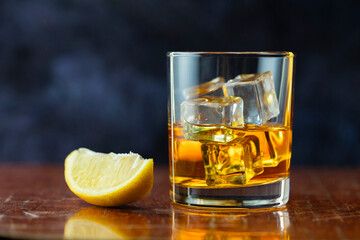 A glass of whiskey with lemon and ice stands on old polished table ion dark background