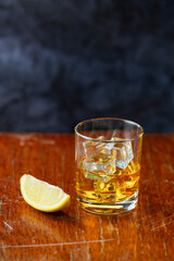 A glass of whiskey with lemon and ice stands on old polished table ion dark background