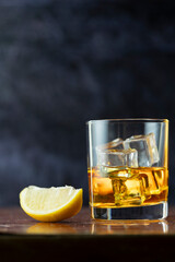 A glass of whiskey with lemon and ice stands on old polished table ion dark background