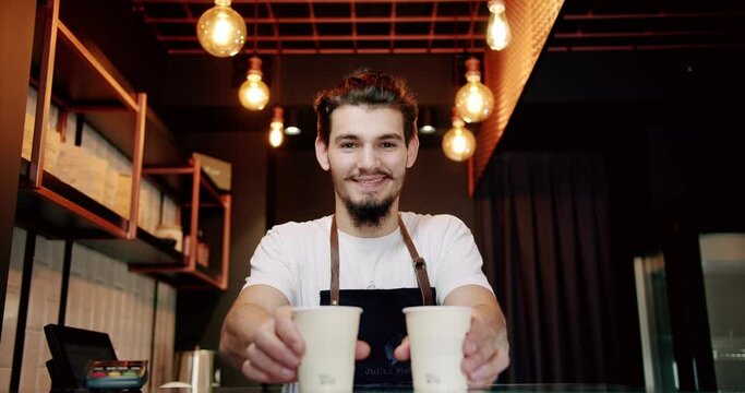Low angle of positive young bearded male barista in white t shirt and apron smiling and looking at camera while serving cups of coffee on counter in modern cafe